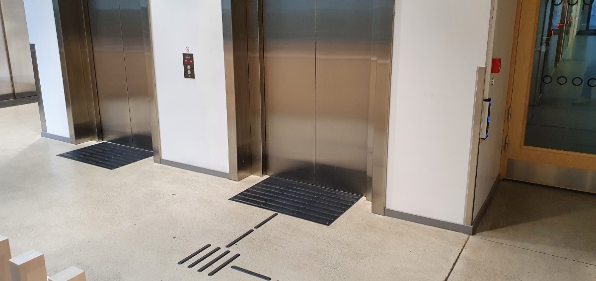 Oznaczenie fakturowe przed windą wraz ze ścieżką naprowadzającą 