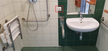 Na zdjęciu widoczna łazienka przystosowana dla osób niepełnosprawnych. Po lewej stronie widoczny prysznic wraz z siedziskiem. Po prawej - umywalka z pochwytami.