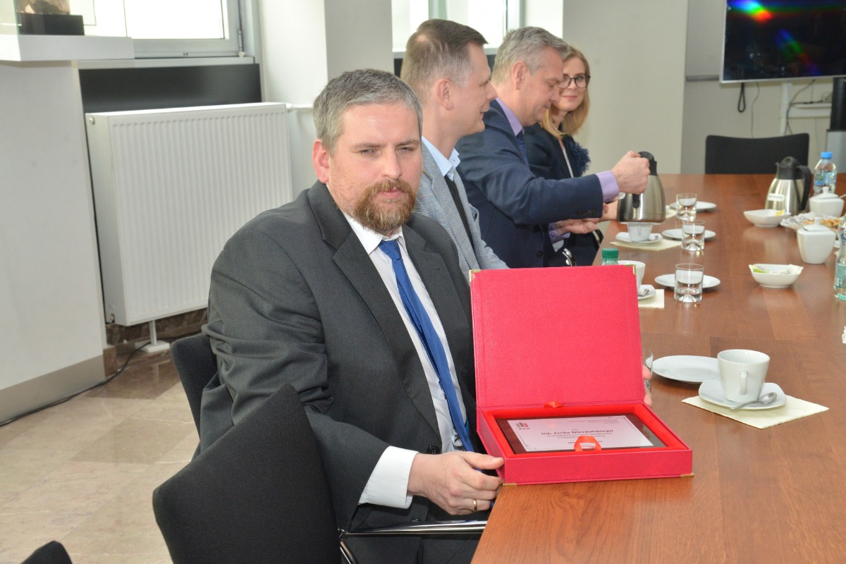 Na zdjęciu siedzący przy stole Pan Jacek Merdalski trzymający w rękach otwarte czerwone pudełko z tabliczką z grawerem w środku. W tle Pan Szczepan Moskwa, Pan Rafał Dańko i Pani Anna Klimczyk 