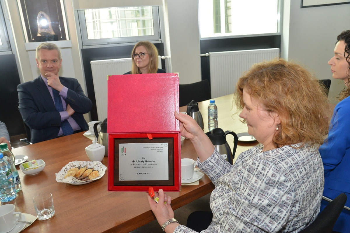 Na zdjęciu siedząca przy stole Pani dr Jolanta Golenia trzymająca w ręce tabliczkę z grawerem w czerwonym pudełku