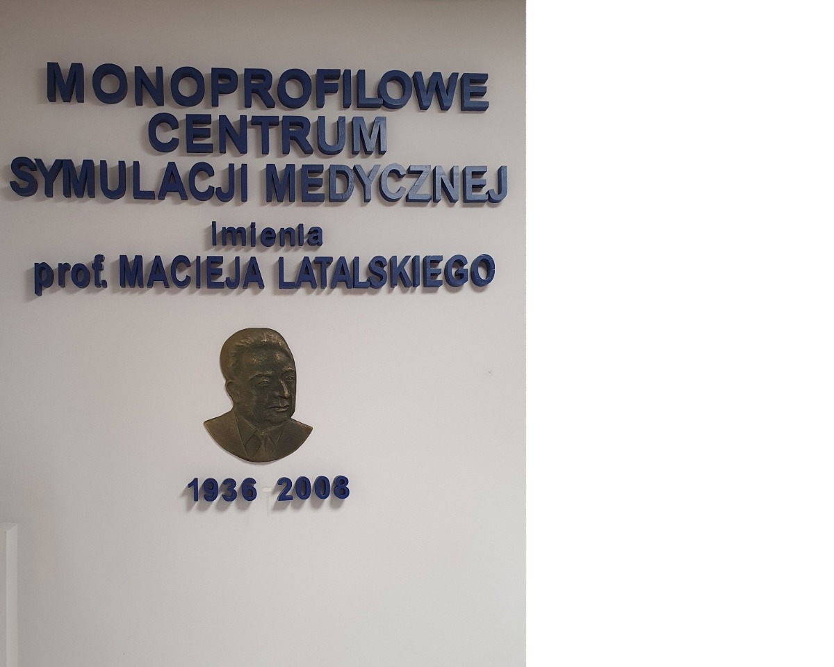 Na ścianie widoczne napis “Monoprofilowe Centrum Symulacji Medycznej imienia prof.. Macieja Latalskiego 1936-2006" i płaskorzeźba portretująca mężczyznę. 