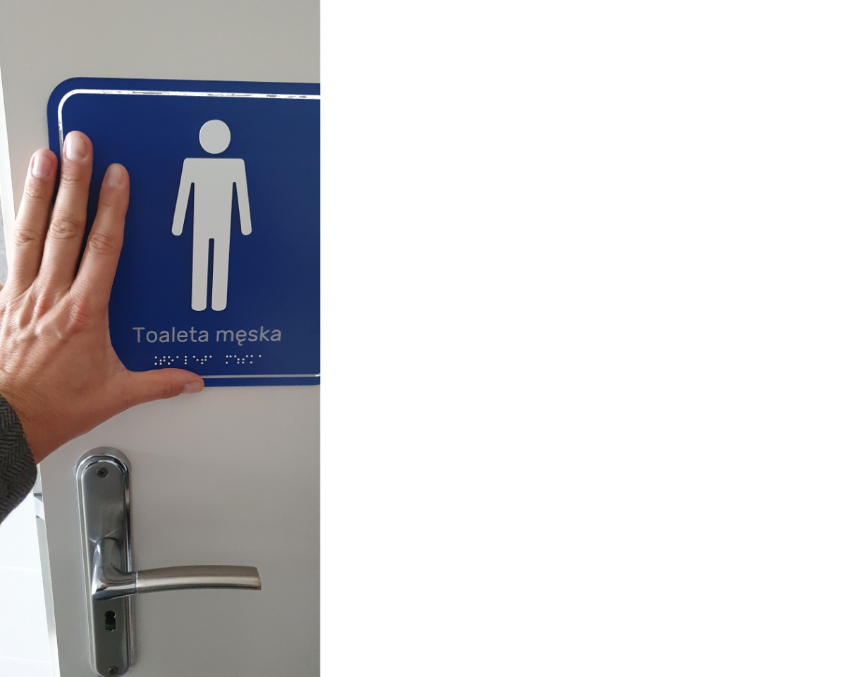 Dłoń przyłożona obok tabliczki informacyjnej WC w kolorach kontrastujących, piśmie wypukłym i alfabecie Breille’a 
